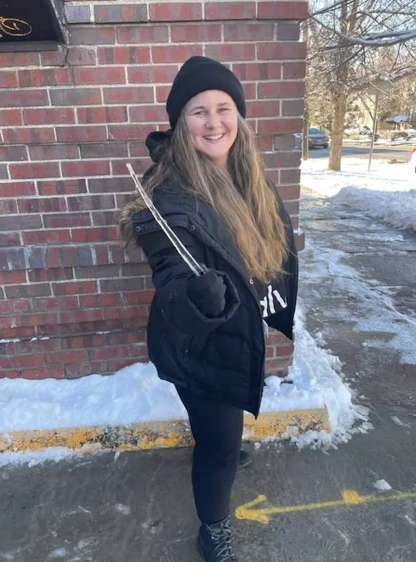 A woman in a black beanie and winter coat holding an icicle by a brick wall