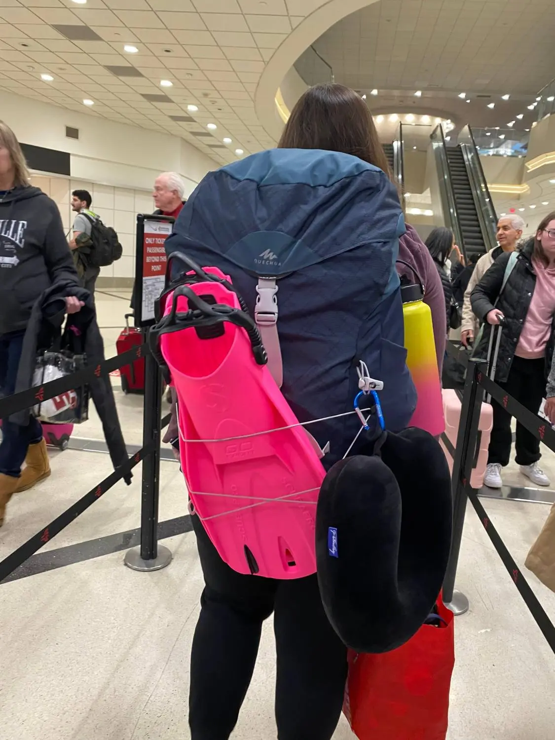 A woman in an airport line with pink dive fins and a neck pillow tied to her backpack