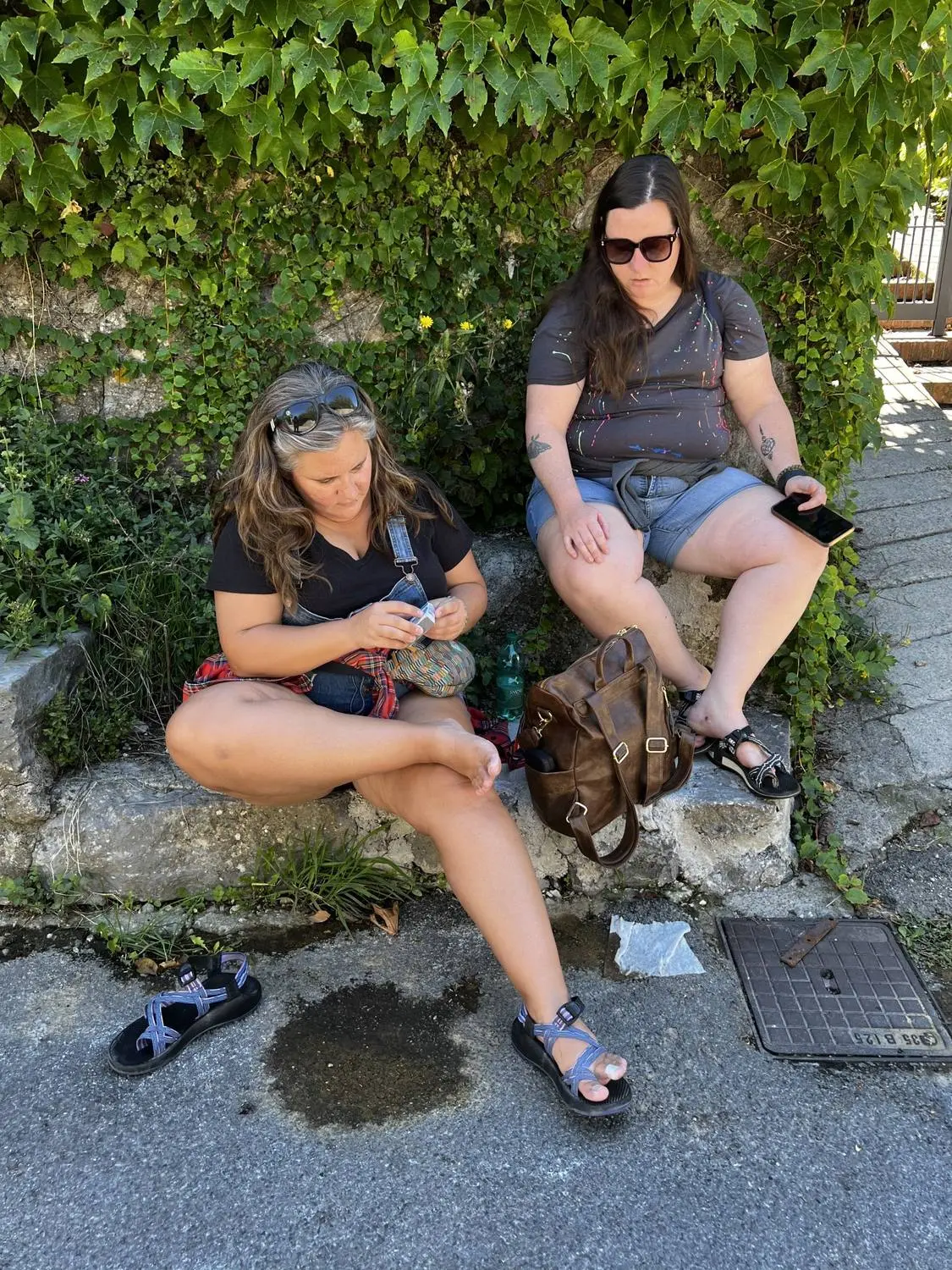 Mariya and Sam sitting on a stone wall by a hedge, checking their phones