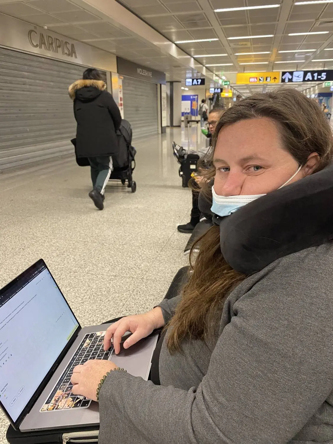 A woman in a neck pillow working on a laptop at an airport gate